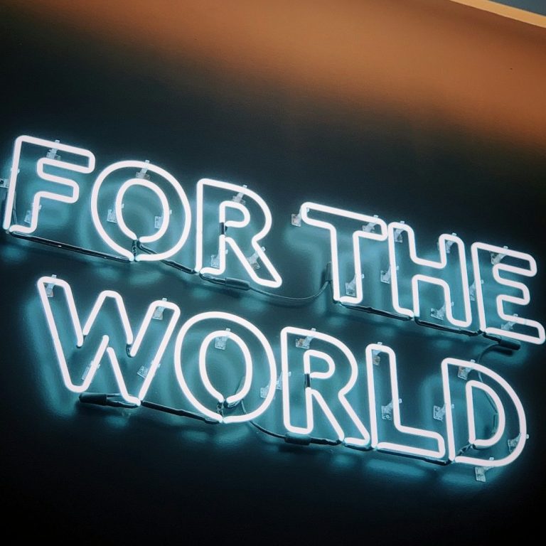 A white neon light in the outline of letters spelling "FOR THE WORLD" is attached to a dark wall